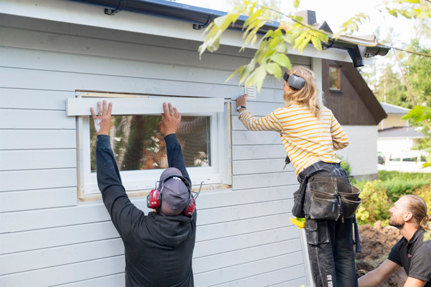 Tre personer hjälps åt med att byta fönster på ett litet trähus.