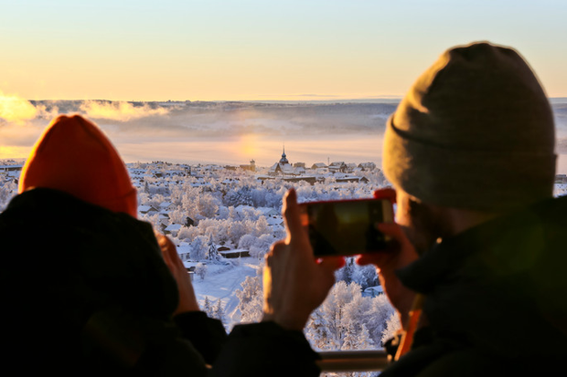 Två personer med mössor i förgrunden. En av dem fotar med mobilen. I bakgrunden syns ett vintrigt Östersund med Rådhusets torn som sticker upp i vinterdiset.