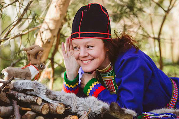 Elin Teilus ligger på en skogstuva iförd samiska kläder, och talar med en sagofigur.