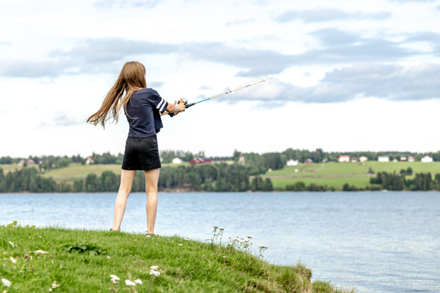 Ung person med långt hår, kjol och t-shirt som fiskar i Storsjön en solig sommardag.