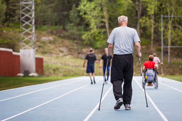 Bild på en idrottsbana där bland annat en äldre man med gåstavar och en rullstolsburen person vistas