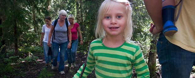 i förgrunden en flicka som är ute i skogen med busig uppsyn. bredvid henne står en förälder med ytterligare ett barn på ryggen. I bakgrunden kommer tre familjemedlemmar till gåendes i skogen längs en stig.