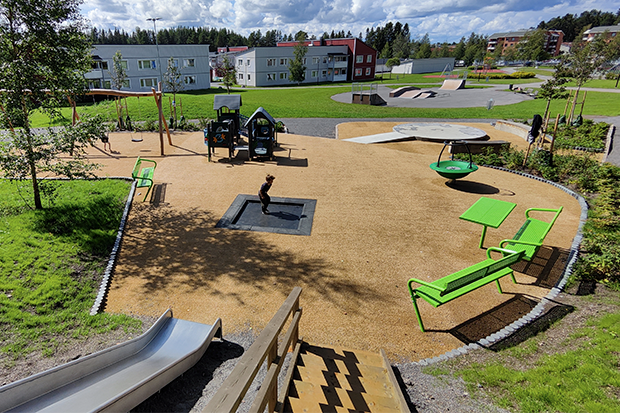 Lekplats med härligt gröna bänkar, lekredskap och ett barn som hoppar på den nedsänkta studsmattan.