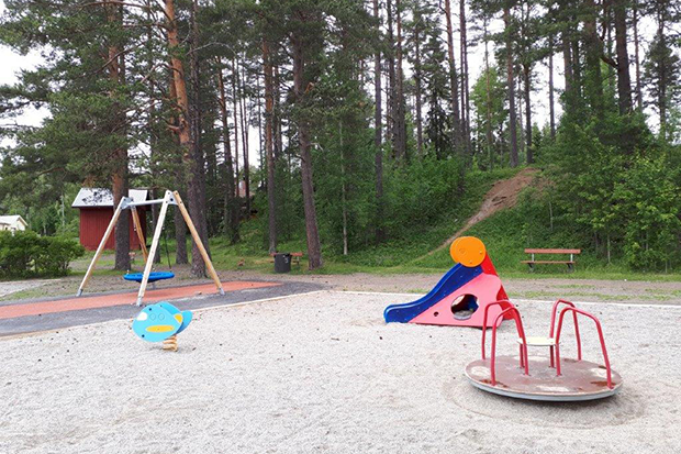 En lekplats med karusell och rutschkana. I bakgrunden syns en kompisgunga.