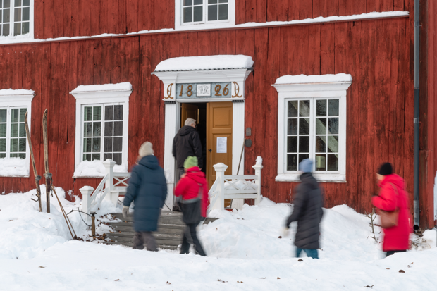 Vinterklädda människor på väg i en rött gammalt hus. Mycket snö