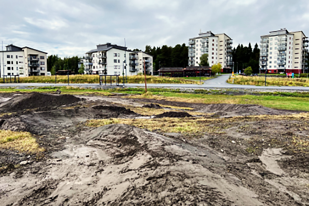 Jordhögar på en öppen yta med gräs, troligen en BMX-bana under uppbyggnad, framför ett bostadsområde med flerbostadshus och gångvägar. I bakgrunden syns träd och en gråmulen himmel.