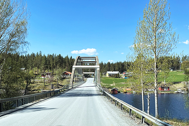 En grusväg leder över en bro omgiven av en vacker landsbygd. På ena sidan av bron syns en sjö med några små hus vid strandkanten. På andra sidan breder en grönskande äng ut sig, kantad av träd och skog. Himlen är klarblå med några vita moln.