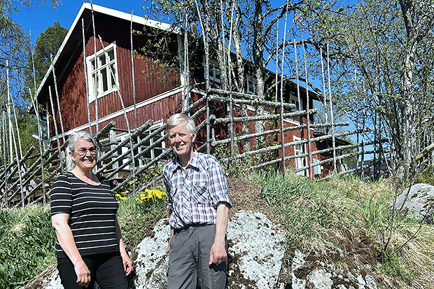 Två personer framför faluröd stuga och gärdesgård i sommarlandskap och klarblå himmel.