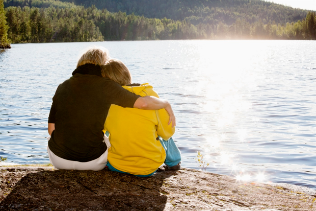 två personer sitter på ett berg. De har ryggen mot kameran och en håller om den andra medans de tittar ut över sjö och skog på andra sidan