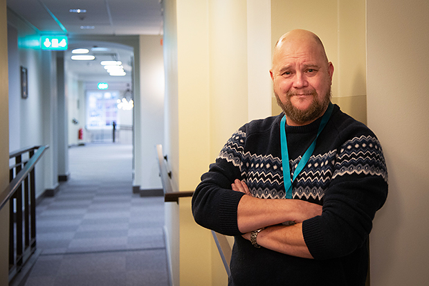 Man med armarna i kors står avslappnat lutad mot en ljus vägg i en kontorskorridor. Han bär en mörk stickad tröja med mönster och ett blågrönt nyckelband runt halsen. I bakgrunden syns en lång korridor med mjuk belysning och flera dörröppningar, vilket skapar djup i bilden.