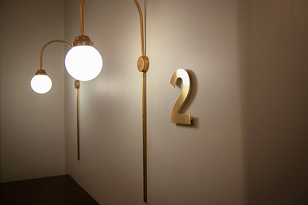 Två vägghängda lampetter i mässing med rundade former och opalvita klotglas lyser upp en ljus vägg. Bredvid den större lampan sitter en guldfärgad siffra 2, fäst direkt på väggen.