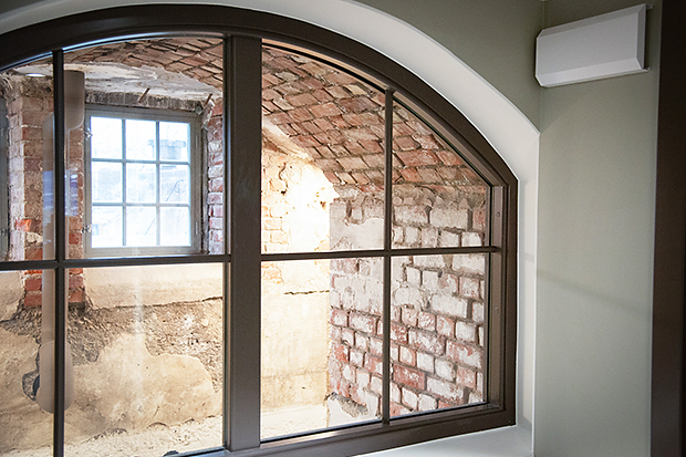 Bågformat innerfönster med mörk spröjsram, som vetter in mot ett äldre valv i röd tegel och putsad sten. Det yttre rummet har en liten rutad fönsteröppning som släpper in dagsljus, vilket belyser de råa tegel- och betongytorna.