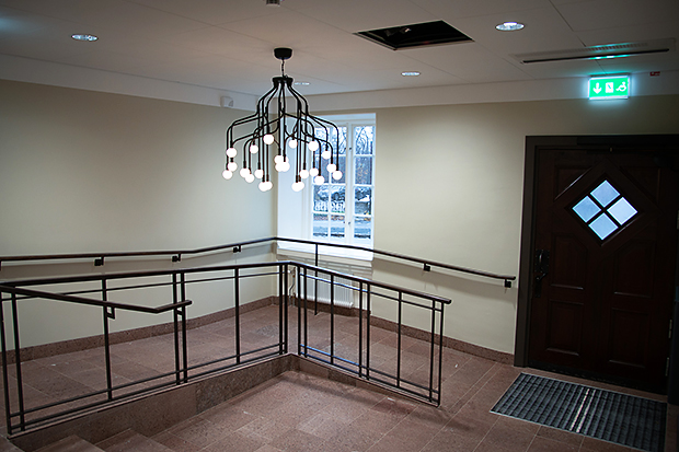 Inomhusentré med rampanpassad nivåskillnad och ledstänger i metall längs väggar och trappor. Golvet är klätt i rödbrun sten, och ett fönster släpper in dagsljus ovanför en radiator. Ett kluster av nakna glödlampor hänger från taket som ljuskrona. Till höger syns en mörk träport med diamantruta och nödutgångsskylt ovanför.