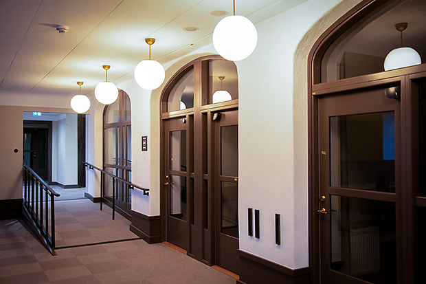 Invändig kontorskorridor med mjuk heltäckningsmatta, rundvälvda dörrar i mörkt trä och glas, och hängande klotformade taklampor i mässing. Längs vänster vägg löper ett ledstångssystem för tillgänglighetsanpassning.
