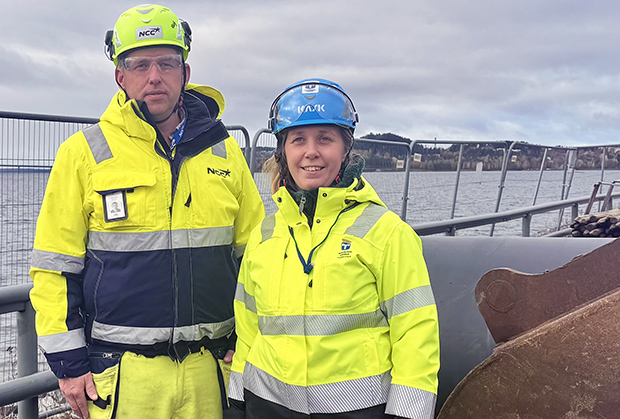 Två personer i gula arbetskläder uppställda för kameran, med Storsjön i bakgrunden.
