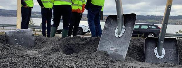 Två spadar i en grushög och i bakgrunden personer med varselvästar pratar med varandra.