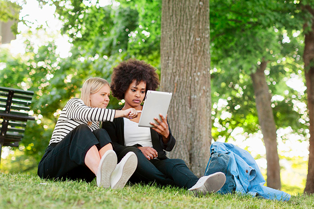 Två kvinnor sitter i gräset under ett träd och tittar i en padda.