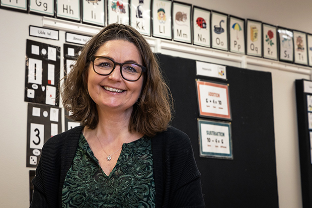 En leende kvinna med glasögon och mörkt axellångt hår står i ett klassrum. Hon bär en grön topp och svart kofta. Bakom henne syns en vägg med alfabetsposters och undervisningsmaterial.