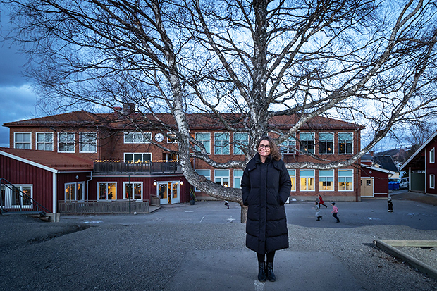 En kvinna klädd i en lång svart vinterjacka står framför en stor björk på en skolgård. I bakgrunden syns en röd skolbyggnad med stora fönster, och några barn leker på gården under en mulen himmel.