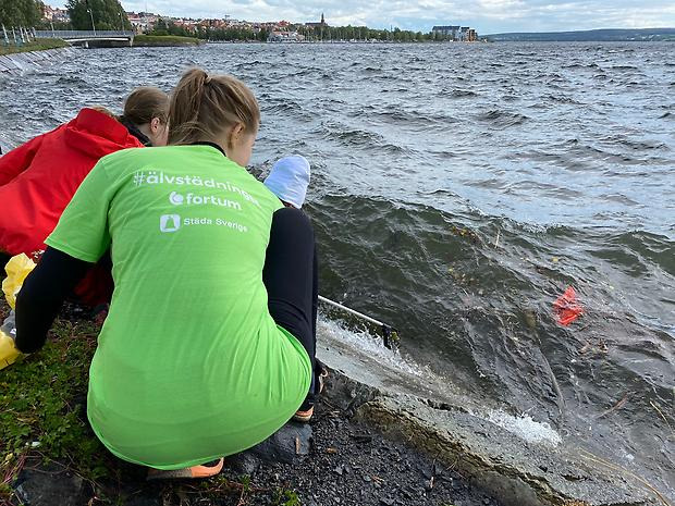 Människor sitter på 
huk vid stranden och håller i ett redskap. I vattnet guppar något rött 
skräp. I horisonten syns Storsjö strand. 