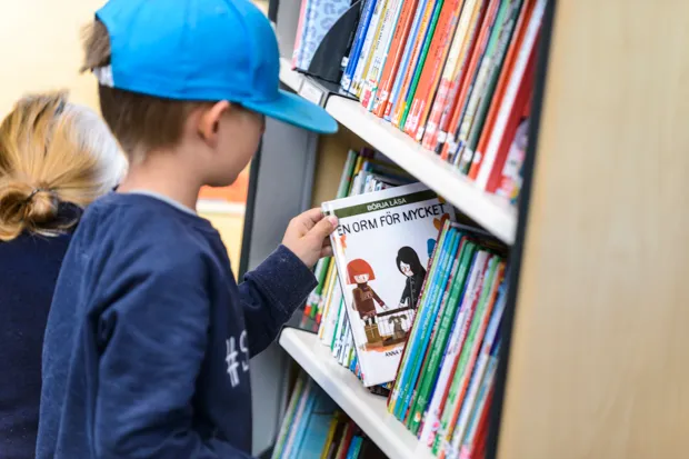 Pojke i blå keps tar fram en bok ur en bokhylla som är fylld med böcker.