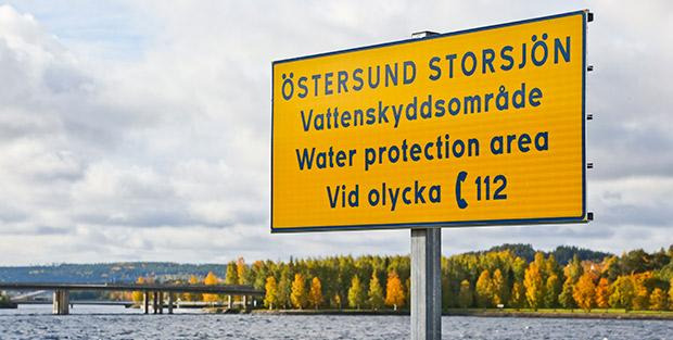 Gul skylt med text om att det är ett vattenskyddsområde. I bakgrunden syns vatten, en bro och träd i höstfärger. 