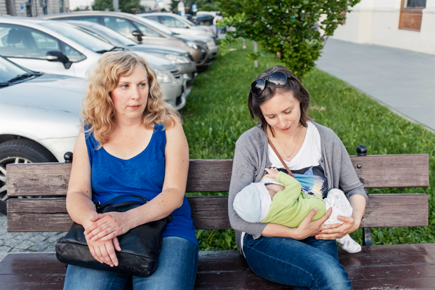 Två kvinnor sitter på en bänk. Den ena tittar på bebisen som hon ammar, den andra tittar i fjärran.