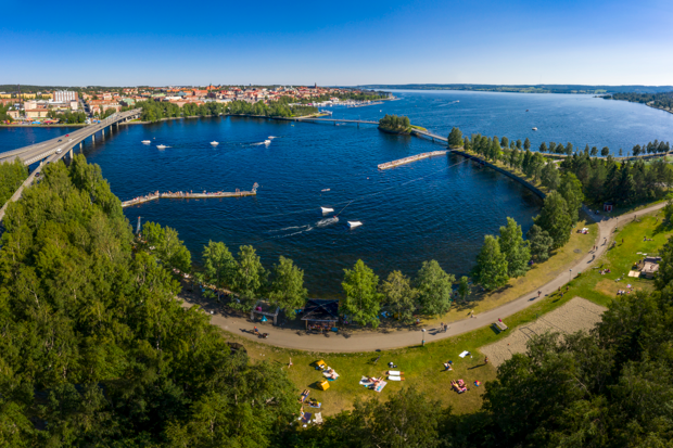 Vy över Surfbukten i Östersund med flera människor som solar, badar och åker wakeboard en solig sommardag. Storsjön glittrar i bakgrunden och vid horisonten syns stadens karaktäristiska skyline.