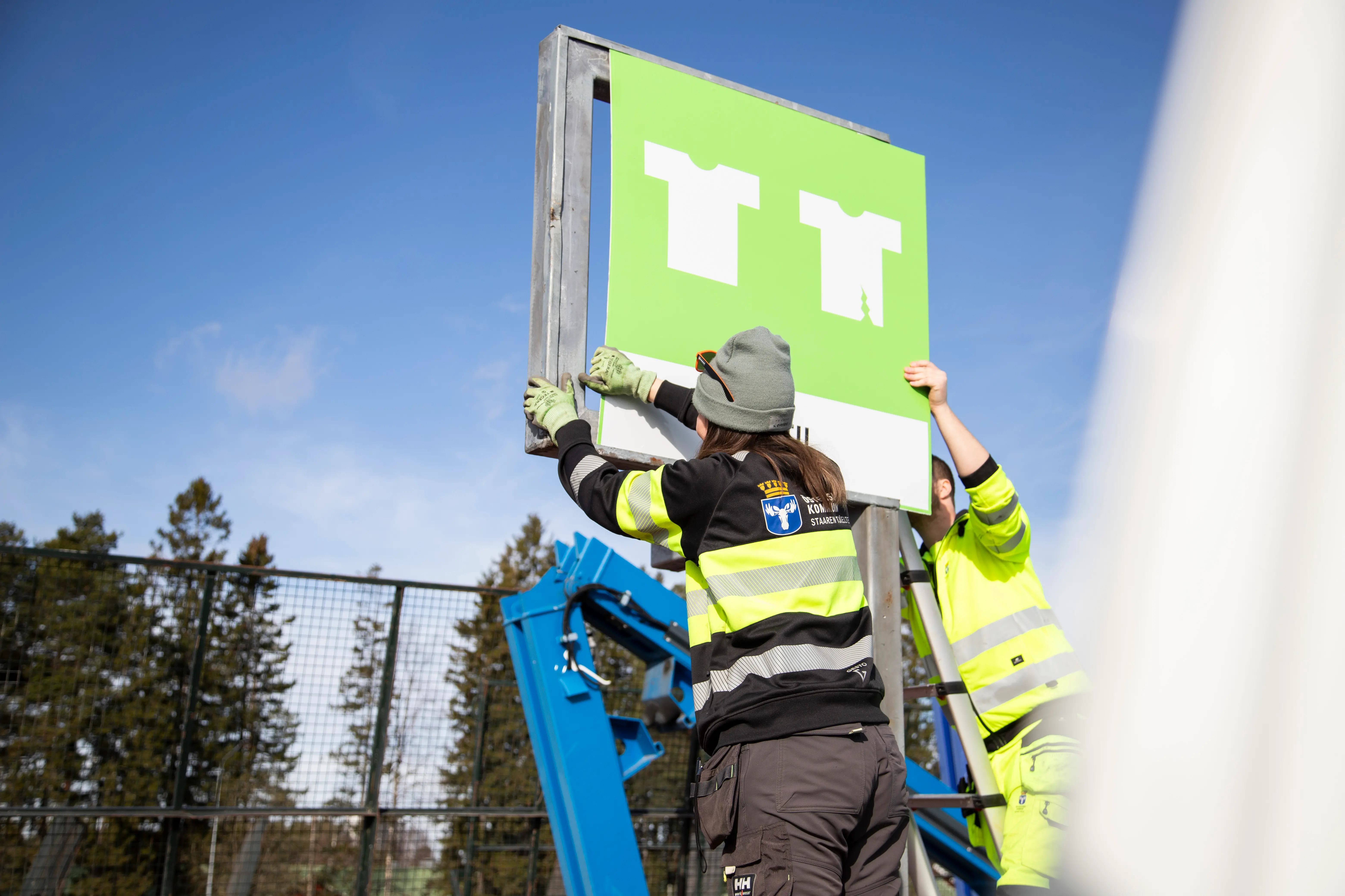 Två personer hjälps åt att sätta upp en grön skylt med texten textil på en stålram.