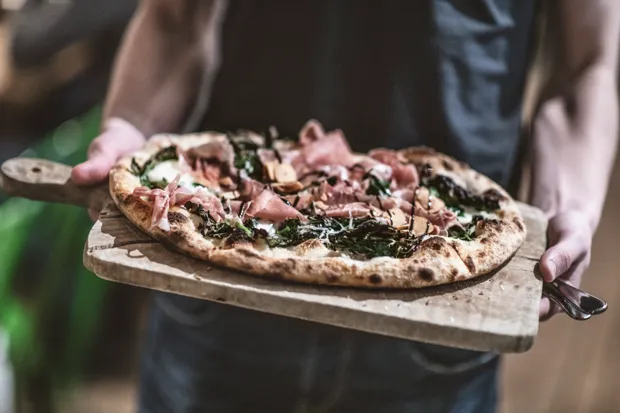 Närbild på en pizza på en skärbräda som hålls av två händer. 