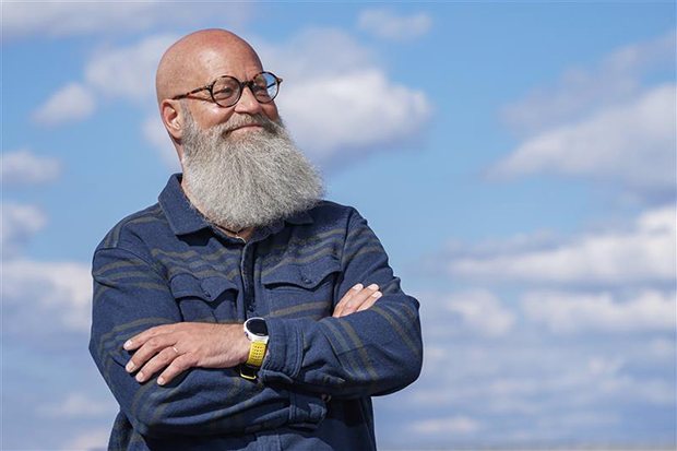 En man med stort grått skägg, och korsade armar, står tryggt och tittar ut mot en utomhusmiljö, med vacker blå himmel och vita moln i bakgrunden.
