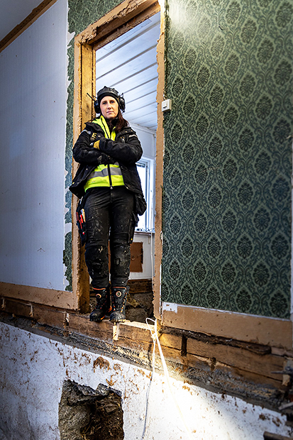 En kvinna i arbetskläder står i en dörröppning på en byggarbetsplats. Hon har armarna i kors och bär skyddsutrustning, inklusive hörselskydd. Rummet har synligt trä och gamla tapeter på väggarna, och golvet är delvis rivet.