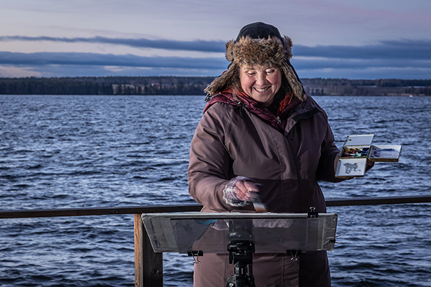 En person målar vid ett staffli utomhus med en sjö i bakgrunden. Hon är klädd i en varm jacka och en pälsfodrad mössa, och håller en färgpalett i ena handen medan hon arbetar med penseln i den andra. Himlen är molnig med ett svagt rosa skimmer vid horisonten.