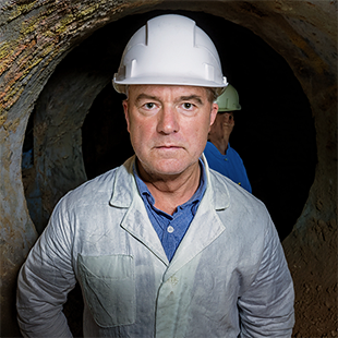 Man i vit skyddshjälm och ljus arbetsrock står framför en tunnelöppning, med en annan person i blå arbetskläder och hjälm i bakgrunden.