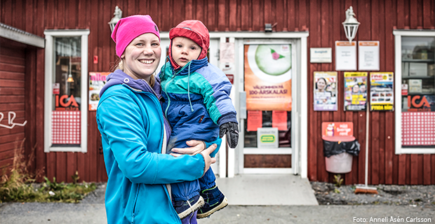 En glad mamma med ett barn i famnen framför ingången till byaffär