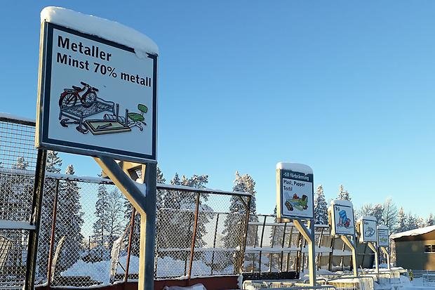 Skyltar och containerlock med snö på överkanterna på återvinningscentralen.