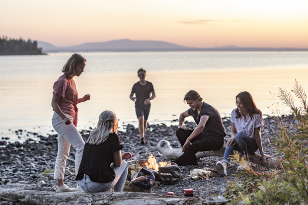 Ett gäng kompisar sitt runt en eld på en strand vid storsjön i skymningen.