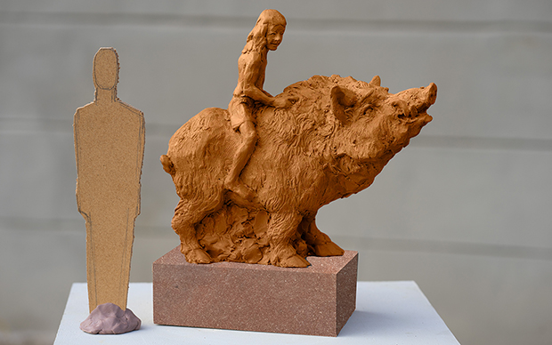 Skulptur i brun lera som föreställer en galt och en pojke som sitter på galtens rygg.