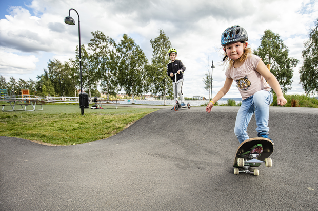 Ett barn åker skateboard och bakom står ett annat barn med en sparkcykel. I bakgrunden syns några björkar och Storsjön.