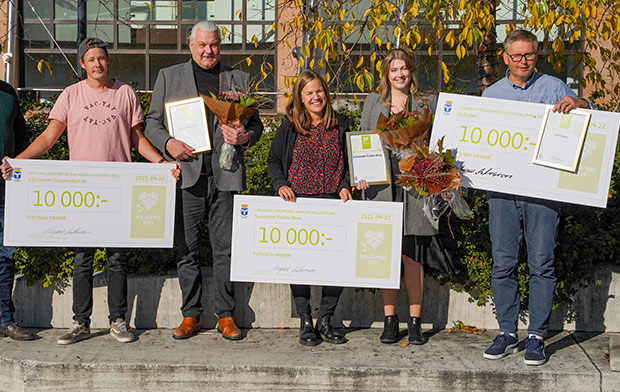 Tre män och två kvinnor står och håller i två stora prischeckar, diplom och blombuketter i solskenet.