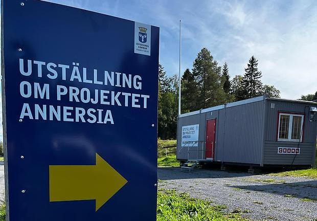 Blå skylt i förgrunden med texten "utställning om projektet Annersia". I bakgrunden en grå barack med röd dörr.