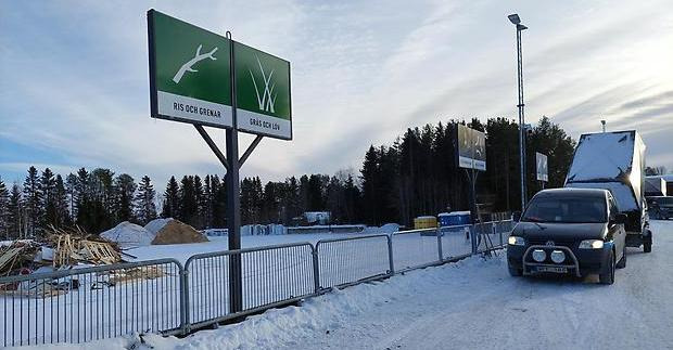 Gröna skyltar, en bil med släp där kåpan är uppfälld, ett staket och en hög med grovavfall. Det är snö på marken