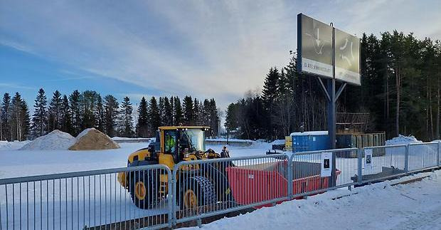 I bakgrunden en hög med träfils. Framför en gul hjullastare som tagit tag i en mindre röd container. Två svarta skyltar på en ramp.