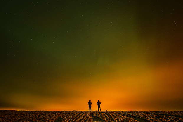 Siluett av två människor på en åker med ett orangefärgat norrsken bakom.