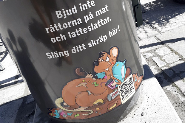 En dekal längst ned på en skräpkorg med en råtta som äter på matrester och texten "Bjud inte råttorna på mat och latteslattar. Släng ditt skräp här!"