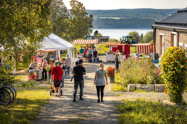 Människor promenerar på en grusväg mot en lantlig marknad med stånd och tält, omgiven av grönska och blommor, med utsikt över en sjö och skog i bakgrunden.