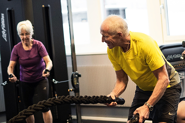 Två äldre personer tränar på ett gym. En man i gul tröja svingar en tung repövning med ett leende på läpparna, medan en kvinna i lila tröja tränar vid en maskin i bakgrunden. De ser fokuserade och engagerade ut i sin träning.
