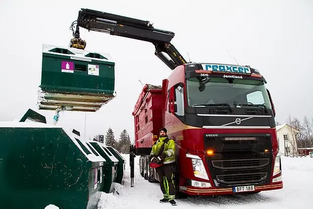 Lastbil lyfter en container med en kran. En person i varselkläder står bredvid lastbilen och styr kranen.