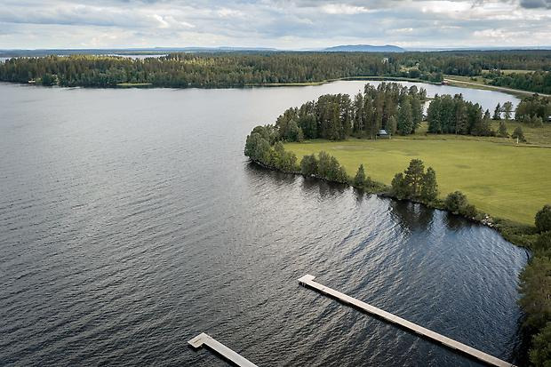 Flygbild över sjö med bryggor i, en udde med träd i sjökanten och skog i horisonten.