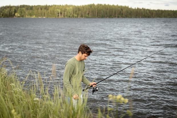 En grönklädd människa med kastspö står vid en sjö. I horisonten är det skog. I förgrunden är det frodigt gräs.